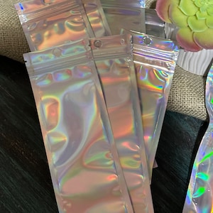 Op de afbeelding: Een verzameling iriserende, holografische zakken met ritssluiting. De zakken zijn rechthoekig met een klein gaatje aan de bovenkant om op te hangen. Het oppervlak reflecteert een regenboog aan kleuren. De zakken zijn waarschijnlijk voor het verpakken van kleine items.