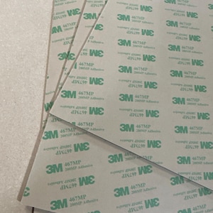 May include: Close-up of several sheets of brown paper with green 3M 467MP 200MP Adhesive labels. The labels are printed with the text "3M 467MP 200MP Adhesive" and "DIN 1297 WE".
