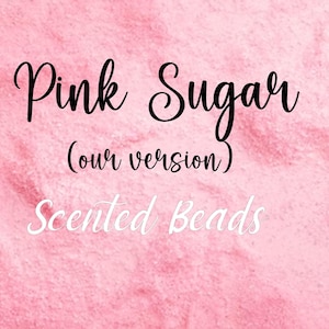 May include: Pink sugar scented beads on a pink background. The text "Pink Sugar (our version)" is written in black cursive font. The text "Scented Beads" is written in white cursive font.