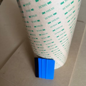 May include: A roll of clear adhesive tape with green text that reads "3M 300LSE" repeated multiple times. A blue and black squeegee is lying on a light brown surface.