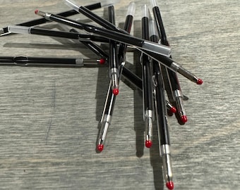 May include: A pile of black and silver pen refills with red tips. The refills are on a light wood surface.