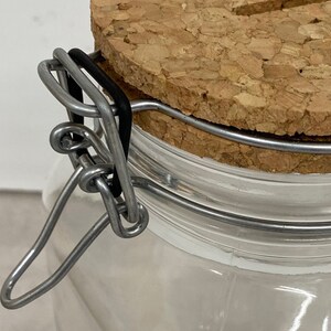 Puede incluir: Un tarro de vidrio con tapa de corcho y cierre de metal. El tarro tiene un cierre de alambre que se asegura con una banda de goma.