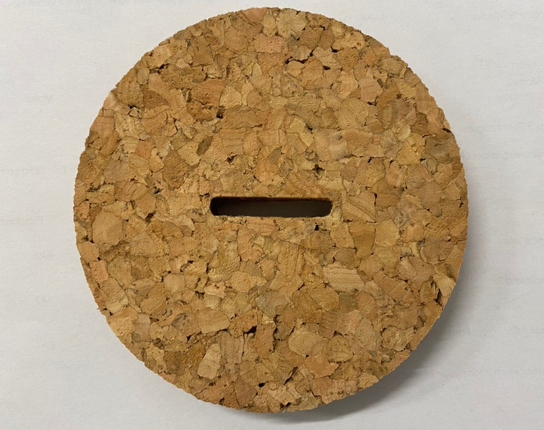 Puede incluir: Un tap&oacute;n redondo de corcho con una ranura rectangular cortada en el centro. El corcho es de color marr&oacute;n claro y tiene una textura rugosa.