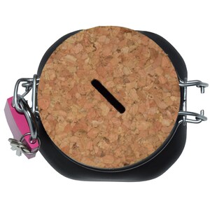 Puede incluir: Un tarro negro con tapa de corcho y un candado rosa. La tapa tiene una ranura para insertar dinero.