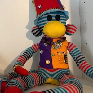 May include: A handmade sock monkey doll with a yellow face, striped body, and a colorful hat. The doll wears a vest with a button and a beaded necklace. The hat is red with gold trim.