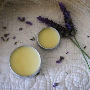 May include: Two small round containers of yellow balm with silver lids, sitting on a white quilted fabric with dried lavender flowers scattered around them.