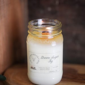 May include: A lit candle in a clear glass jar with a white wax interior and a wooden wick. The jar has a label that reads "Brown Sugar Fig" and "Alvarez River Farms." The candle is set on a wooden surface with a dark brown background.