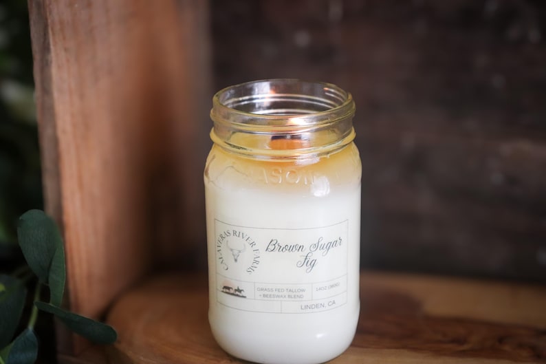 May include: A lit candle in a clear glass jar with a white wax interior and a wooden wick. The jar has a label that reads "Brown Sugar Fig" and "Alvarez River Farms." The candle is set on a wooden surface with a dark brown background.