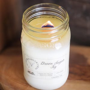May include: A lit candle in a clear glass jar. The candle has a white base and a yellow top, with a visible flame. The jar has a label that reads "Brown Sugar Fig".