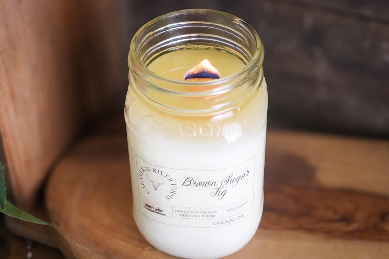 May include: A lit candle in a clear glass jar. The candle has a white base and a yellow top, with a visible flame. The jar has a label that reads "Brown Sugar Fig".