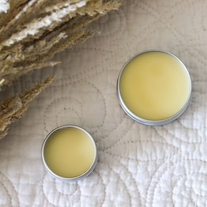 May include: Two round metal tins with lids, containing a yellow balm or salve. The tins are on a white quilted fabric background.