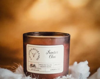 May include: A brown glass candle jar with a white label that reads "Frontier Chai." The label also includes the text "Grass Fed Tallow + Beeswax Blend" and "Smoky pine and cinnamon spice meet vanilla." The candle is 14 oz.
