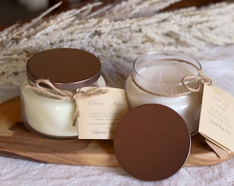 May include: Two glass jar candles with bronze lids sit on a wooden tray. One candle has a Citrus-Agave scent tag, the other a White Oak Vanilla scent tag. Both are tied with twine. The candles are surrounded by decorative dried plants.