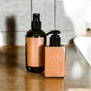 May include: Two amber glass bottles with black pump tops and labels that read "Kojic Acid Niacinamide Brightening Wash". The smaller bottle has the text "With Niacinamide, Kojic Acid, Organic Cucumber Extract, Organic Oat".