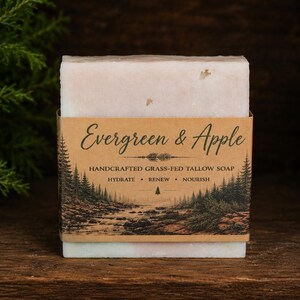 May include: A rectangular bar of soap wrapped in a brown paper label featuring the text Evergreen & Apple, Handcrafted Grass-Fed Tallow Soap, Hydrate, Renew, Nourish, and an illustration of a forest landscape with pine trees and a stream.