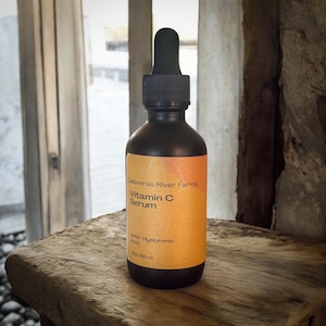 May include: A dark brown glass bottle of Vitamin C Serum with a dropper. The label is orange with the text "Calaveras River Farms Vitamin C Serum" and "With Hyaluronic Acid". The bottle contains 2 fl oz (60 ml).