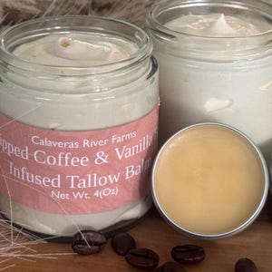 May include: Two glass jars and two small tins of whipped balm. The jars have pink labels that read "Whipped Coffee & Vanilla Infused Tallow Balm." The small tins contain a golden balm. Coffee beans are scattered around.