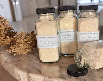 Könnte beinhalten: Vier Glasbehälter gefüllt mit einem weißen Badezusatzprodukt. Die Behälter haben schwarze Deckel und Etiketten mit der Aufschrift "Calaveras River Farms" und "Soothe Bath Soak".