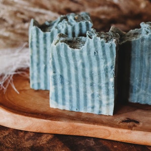 May include: Four rectangular bars of handmade soap with a blue and white striped design are arranged on a wooden tray. The soap has a textured, uneven top. The tray is made of light brown wood. Decorative feathers are in the background.