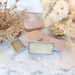 May include: Four small, rectangular tins with lids. Three of the tins are closed and one is open, revealing a light green balm inside. The tins are on a white marble surface.