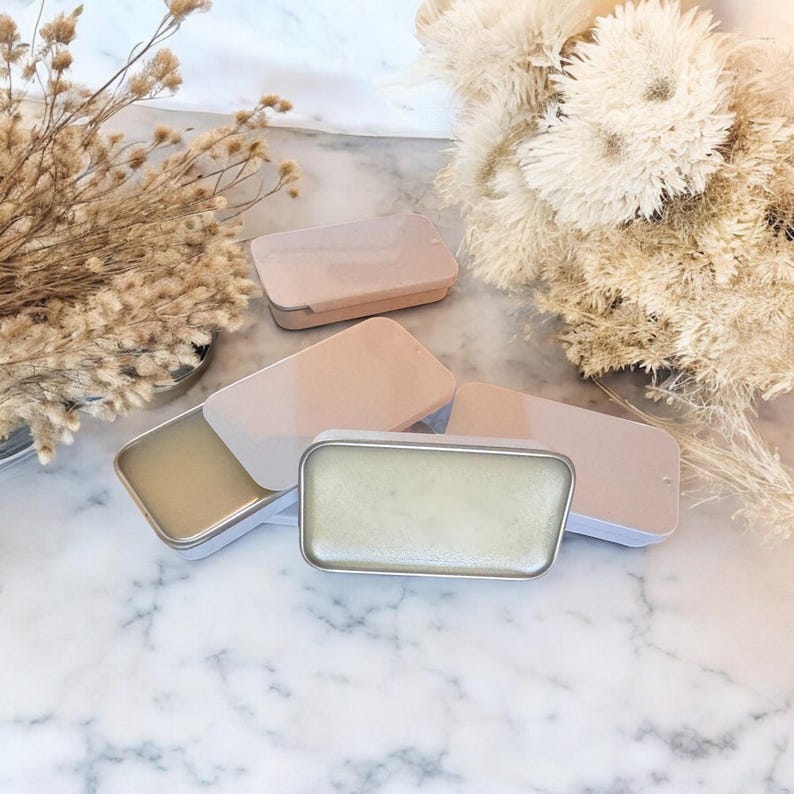 May include: Four small, rectangular tins with lids. Three of the tins are closed and one is open, revealing a light green balm inside. The tins are on a white marble surface.