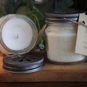 May include: Two glass jar candles with metal lids and a tag that reads "Red Berry Oasis". One candle is open, revealing the white wax and wick. The other candle is closed. The candles sit on a wooden surface.