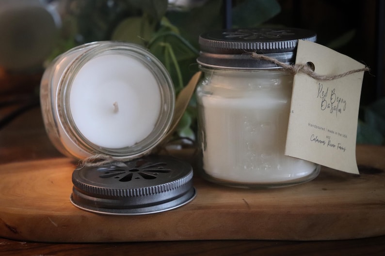 May include: Two glass jar candles with metal lids and a tag that reads "Red Berry Oasis". One candle is open, revealing the white wax and wick. The other candle is closed. The candles sit on a wooden surface.