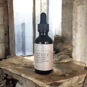 May include: A dark brown glass bottle of serum with a black dropper. The label reads "Brightening Kojic Acid & Hyaluronic Acid with Niacinamide Serum" from Calaveras River Farms. The bottle is sitting on a stone surface.