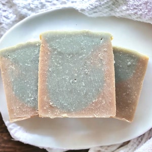 Puede incluir: Tres jabones artesanales sobre un plato blanco. Los jabones tienen un diseño en capas con un centro azul claro, un borde marrón rosado y un exterior marrón claro. Los jabones están colocados sobre un plato blanco con un paño blanco en el fondo.