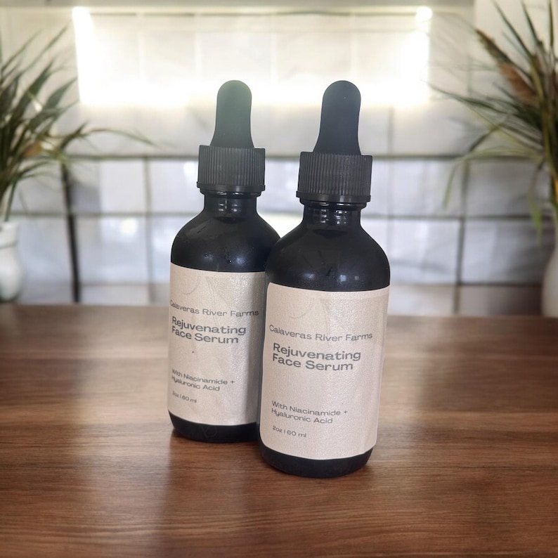 Puede incluir: Dos botellas de vidrio negro con goteros que contienen un suero facial. Las botellas est&aacute;n etiquetadas como "Calaveras River Farms Rejuvenating Face Serum" y enumeran los ingredientes como "With Niacinamide + Hyaluronic Acid".