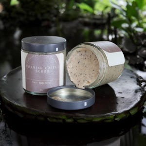 May include: Two glass jars of foaming coffee scrub, one with the lid off, showing the brown scrub inside. The jar has a label that reads "Foaming Coffee Scrub, Face + Body Scrub, 4 oz."