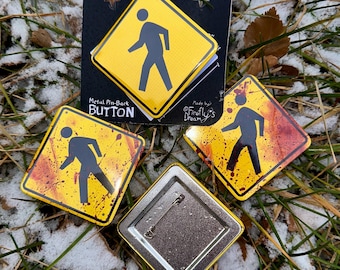 May include: A selection of metal pin-back buttons with a pedestrian crossing sign design. The buttons are yellow with a black pedestrian figure and a red splatter effect. One button is on a black backing with the text "Metal Pin-Back Button".