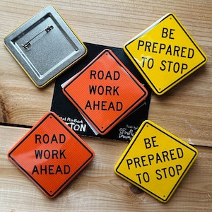 May include: A collection of metal pin-back buttons with road sign designs. The buttons feature the phrases "Road Work Ahead" in orange and "Be Prepared to Stop" in yellow. One button is a square with a pin back.