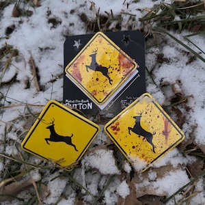 May include: Yellow metal fin-back buttons featuring a black deer silhouette on a yellow background, styled like a road sign. The buttons have a distressed appearance with red splatter details. The buttons are on snow and grass.