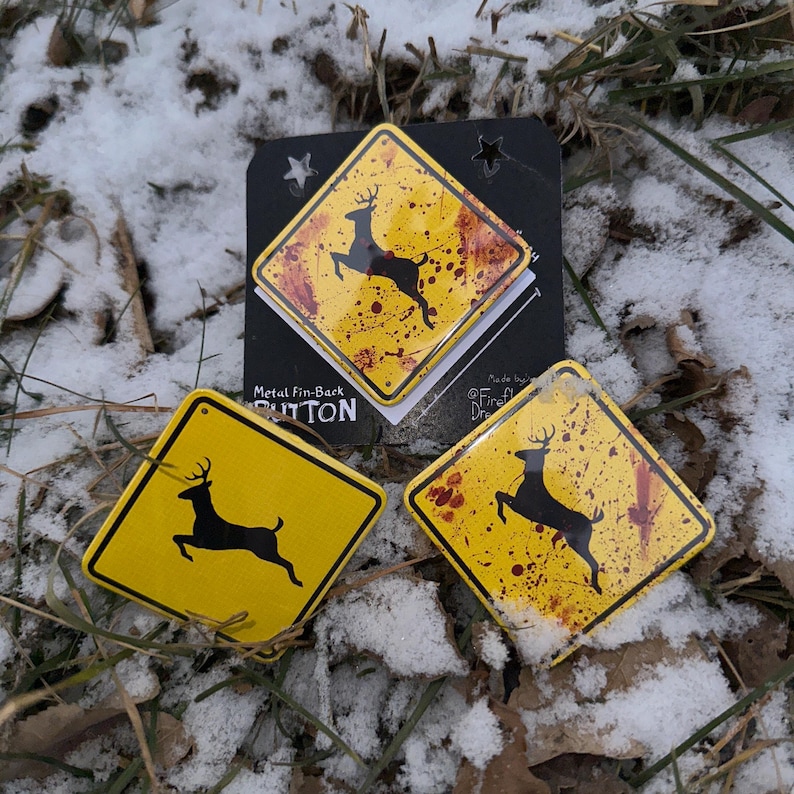May include: Yellow metal fin-back buttons featuring a black deer silhouette on a yellow background, styled like a road sign. The buttons have a distressed appearance with red splatter details. The buttons are on snow and grass.