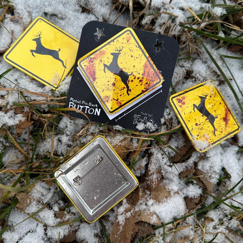 May include: A selection of metal pin-back buttons featuring a yellow and black deer crossing sign design. The buttons are square and have a safety pin on the reverse. The text "Metal Pin-Back Button" is visible on the packaging.