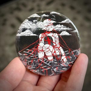 Op de afbeelding: Een zwart-witte knoop met een rood touwontwerp. Het ontwerp toont een persoon die zit met rood touw om zich heen gewikkeld. De achtergrond is zwart met witte wolken.