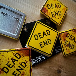 May include: A collection of yellow and black "Dead End" metal pin-back buttons with a distressed, rusty appearance. The buttons are square with a diamond shape and feature the words "DEAD END" in bold black letters. Some buttons have a blood splatter design.
