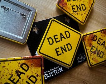 May include: Several yellow and black "Dead End" metal pin-back buttons with a distressed, rusty look. The square buttons feature a diamond shape and the words "DEAD END" in black. Some have a blood splatter design, and one has a metal pin back.