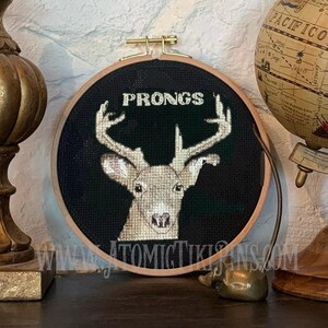 May include: A cross-stitch embroidery hoop featuring a deer with the word "PRONGS" above its antlers. The deer is stitched in shades of brown and white against a black background. The hoop is set in a wooden frame.