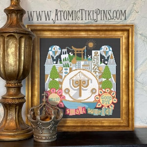 May include: Framed cross-stitch art with a gold frame. The artwork features a colorful depiction of a whimsical cityscape with hot air balloons, trees, and the words "small world." The text "HELLO" and "GOOD BYE" are also included.