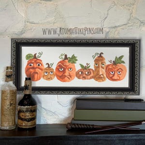 Puede incluir: Un bordado enmarcado que representa seis calabazas naranjas con diversas expresiones faciales y hojas verdes. La obra de arte enmarcada en negro se coloca sobre una tela blanca. La dirección del sitio web www.atomictikipins.com es visible. Tema de Halloween.
