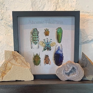 May include: A framed cross-stitch design featuring a collection of colorful beetles, including a green beetle, a blue beetle, and a brown beetle. The design is titled "Atomic Tiki Pins".
