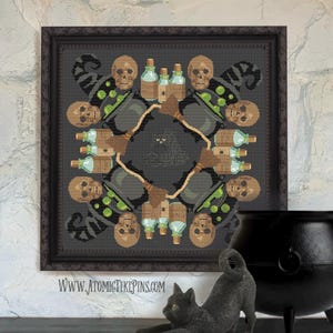 May include: Framed cross-stitch art with a dark grey background featuring a circular design of skulls, witch hats, potion bottles, cauldrons, and brooms. A black cat statue and a cauldron are in the foreground. The website address www.atomictikipins.com is at the bottom.