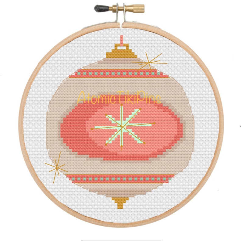 May include: A cross-stitch design of a retro Christmas ornament in a wooden embroidery hoop. The ornament features a light beige background with a red oval center and a green and gold starburst design. The text "Atomic Tiki Pins" is visible.
