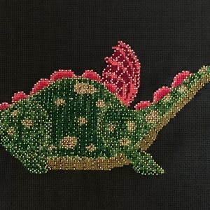 May include: An embroidered dragon in green and red with a small brown bucket in its mouth. The dragon is stitched onto a black fabric background.