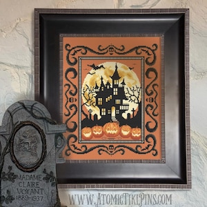 May include: Framed Halloween cross-stitch art depicting a haunted house silhouette against a full moon. The design includes jack-o'-lanterns, bats, and a decorative border. The frame is dark brown, and the artwork is displayed with a tombstone prop.