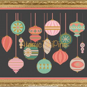 May include: A framed cross-stitch artwork featuring a collection of retro Christmas ornaments in shades of pink, teal, and gold against a dark gray background. The text "Atomic TriPings" is visible. The ornaments have various geometric and starburst designs.