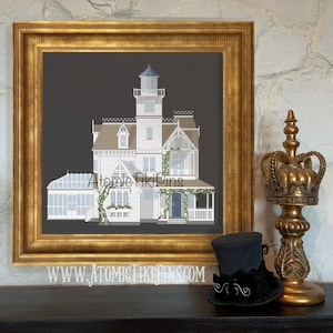 May include: A framed cross-stitch of a white Victorian house with a blue door and a lighthouse-style tower. The frame is gold. A black top hat with a decorative bow and a gold crown-shaped candle holder are on a dark surface.