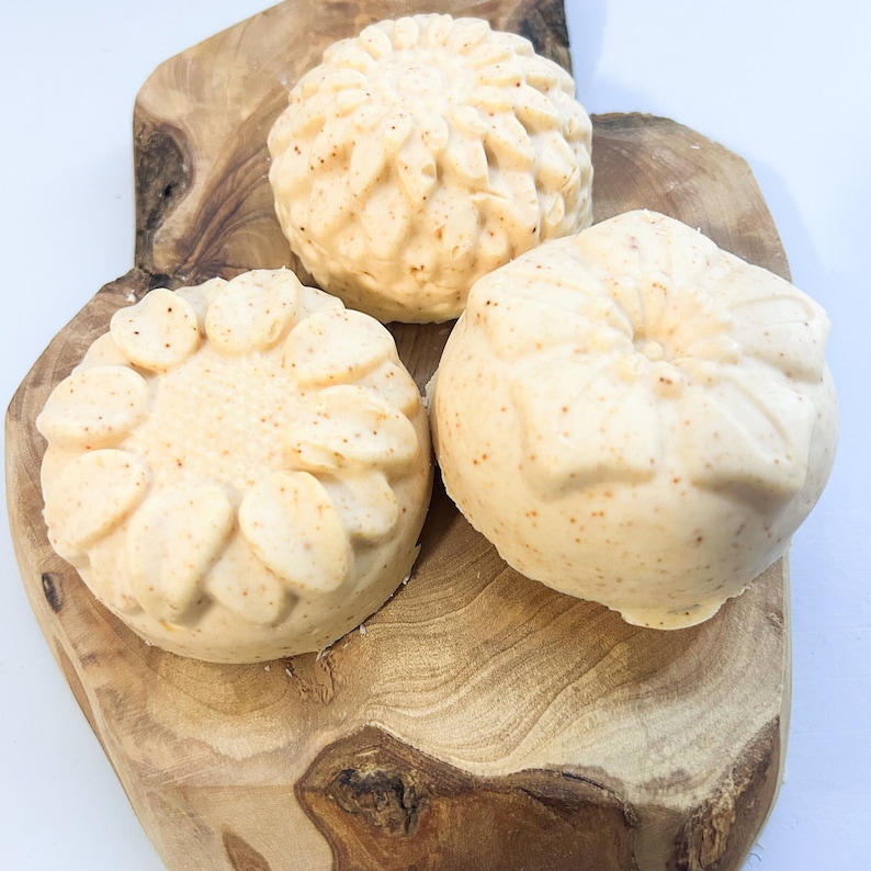 Peut inclure: Trois savons ronds de couleur blanc cass&eacute; avec un motif floral sur une surface en bois. Les savons ont une texture mouchet&eacute;e.